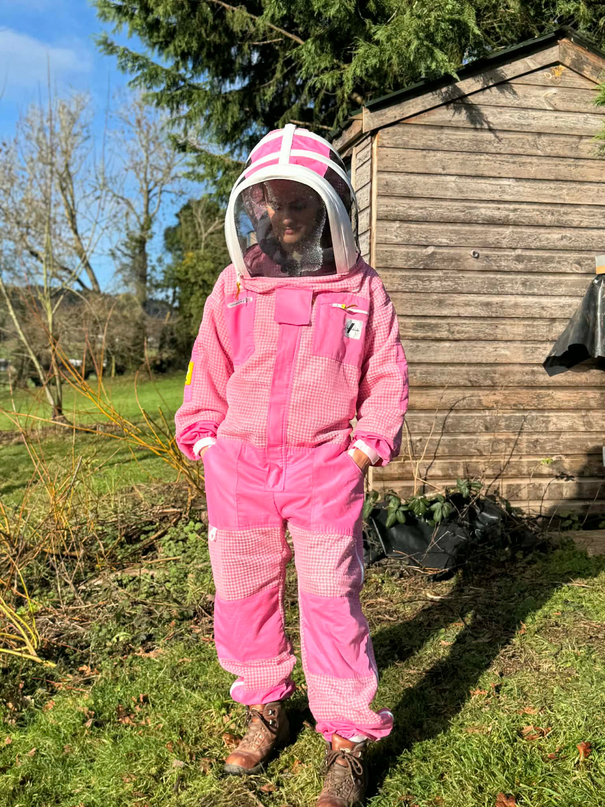 Person wearing a pink beekeeping suit standing outdoors near a wooden shed.